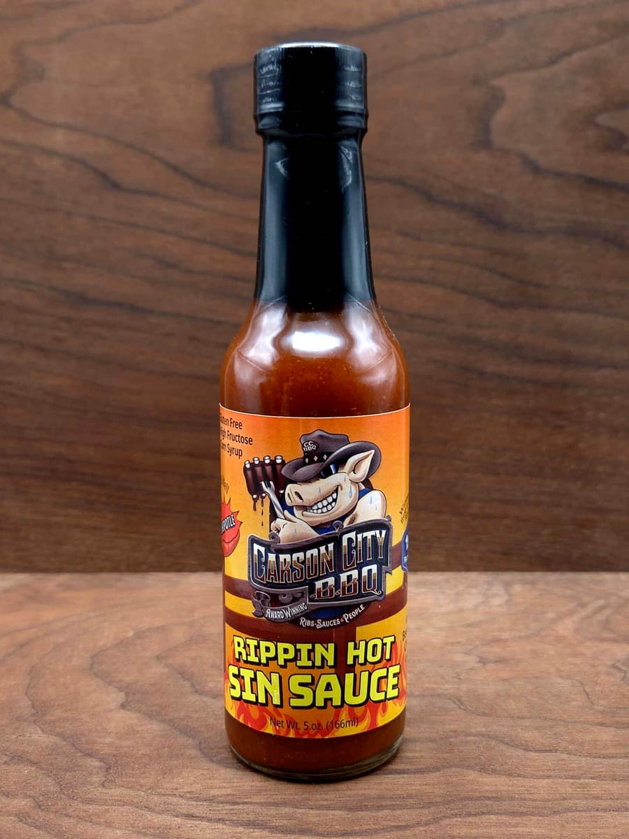 A tall five-ounce bottle of Carson City BBQ Chipotle Sin Sauce with a black cap, labelled Rippin Hot Sin Sauce, standing on a dark wood surface.