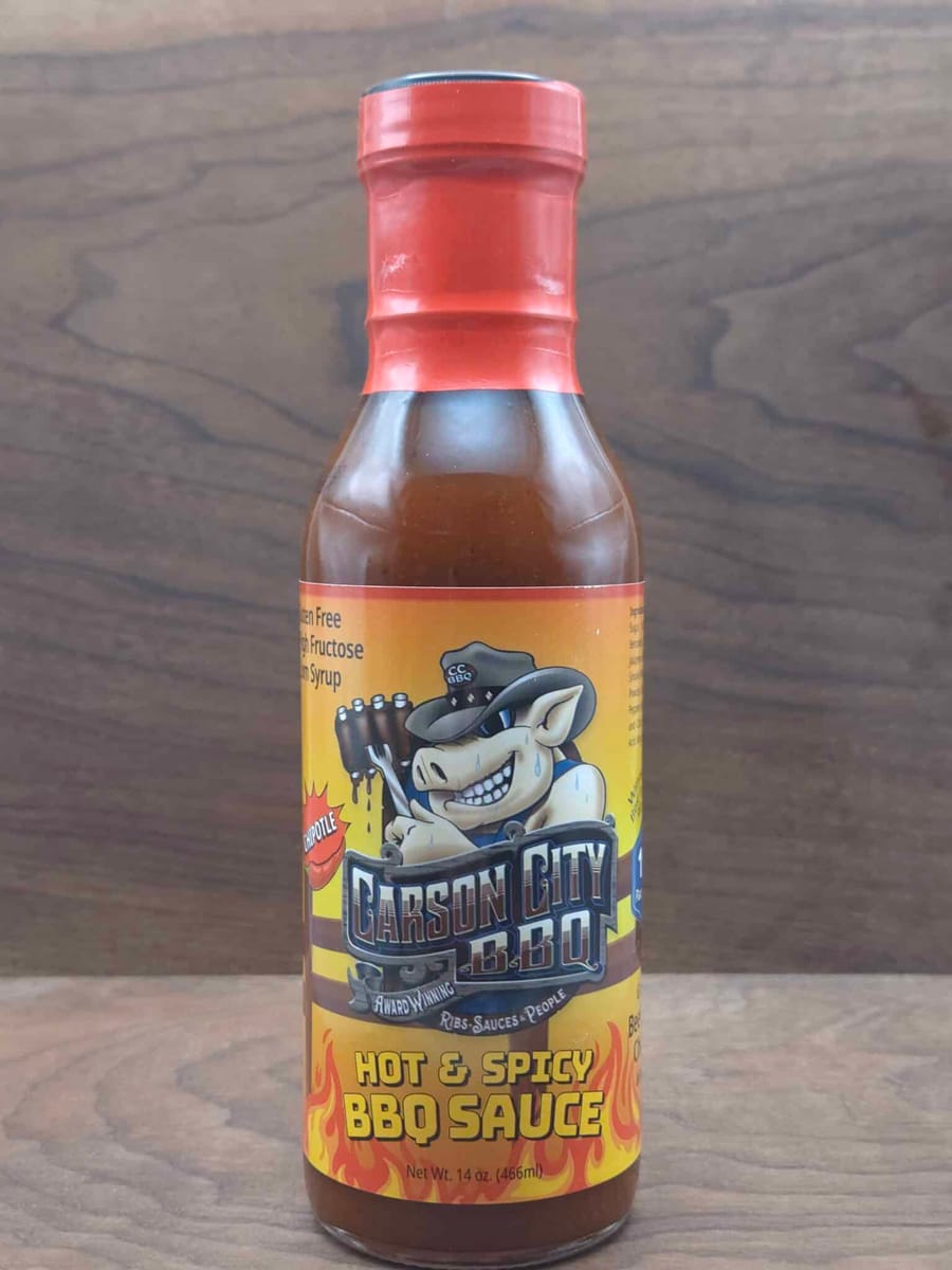 A bottle of Carson City BBQ Hot & Spicy BBQ Sauce with a red cap, standing on a dark wood surface. The label shows the Carson City BBQ pig mascot in a cowboy hat above flames.