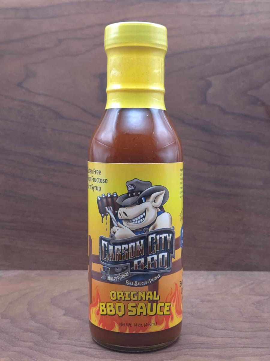 A bottle of Carson City BBQ Original BBQ Sauce with a yellow cap, standing on a dark wood surface. The label shows the Carson City BBQ pig mascot in a cowboy hat above flames.