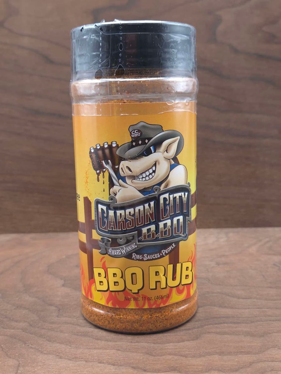 A shaker of Carson City BBQ all-purpose BBQ Rub on a dark wood surface, the coarse orange-red seasoning visible through the clear lid.